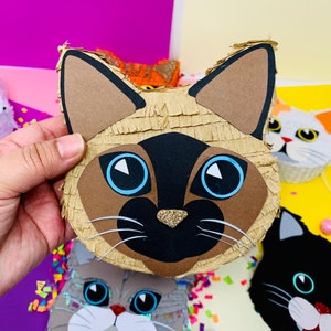 May include: A collection of cat-shaped pi&ntilde;atas. The primary pi&ntilde;ata is a Siamese cat with a brown face, black nose, and blue eyes. Other cat pi&ntilde;atas include a gray cat, a black cat, and an orange cat. The pi&ntilde;atas are decorated with glitter and confetti.