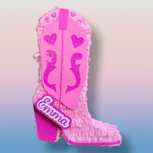 Può includere: Una pignatta a forma di stivale da cowboy rosa con un design decorativo. Lo stivale presenta un tacco rosa, un gambale rosa con ritagli a forma di cuore e un'etichetta con la scritta "Emma". La pignatta è fatta di carta frangiata.