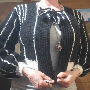 Könnte beinhalten: Schwarz-weiß gestreifter, verkürzter Cardigan mit einer weißen gehäkelten Schleife am Hals. Der Cardigan hat lange Ärmel und einen weißen Saum am unteren Rand.