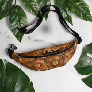 Puede incluir: Riñonera marrón con un diseño de caparazón de tortuga y una correa negra. La riñonera tiene una pequeña figura de tortuga en la parte delantera. Tiene cremallera y hebilla negras.