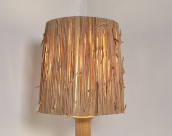 May include: A wooden table lamp with a natural woven shade. The shade is made of thin strips of natural material, possibly straw or bamboo. The lamp is lit and casting a warm glow.