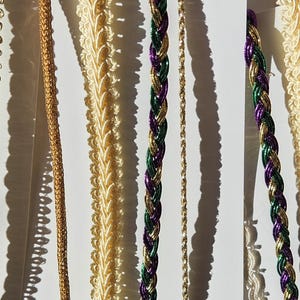 May include: An assortment of decorative trims in various styles and colors. The trims include braided, beaded, and scalloped designs in gold, cream, purple, and green. The trims are arranged vertically against a white background.