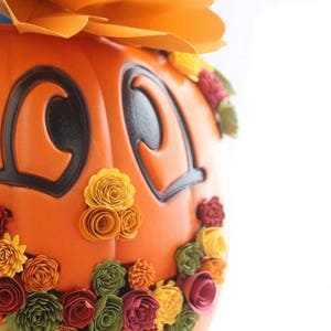 May include: Orange pumpkin with a smiling face, decorated with fall-colored paper flowers. The pumpkin has black eyes and a wide, toothy grin.