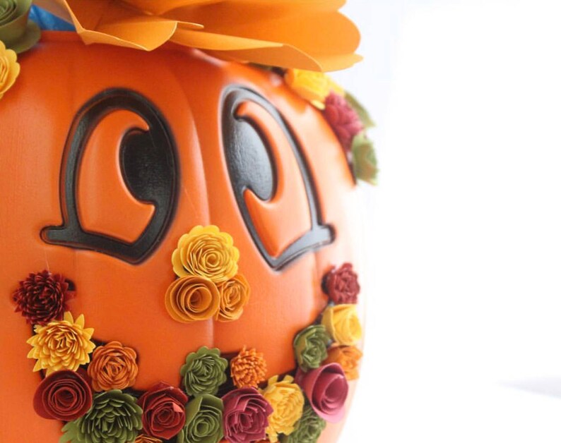 May include: Orange pumpkin with a smiling face, decorated with fall-colored paper flowers. The pumpkin has black eyes and a wide, toothy grin.