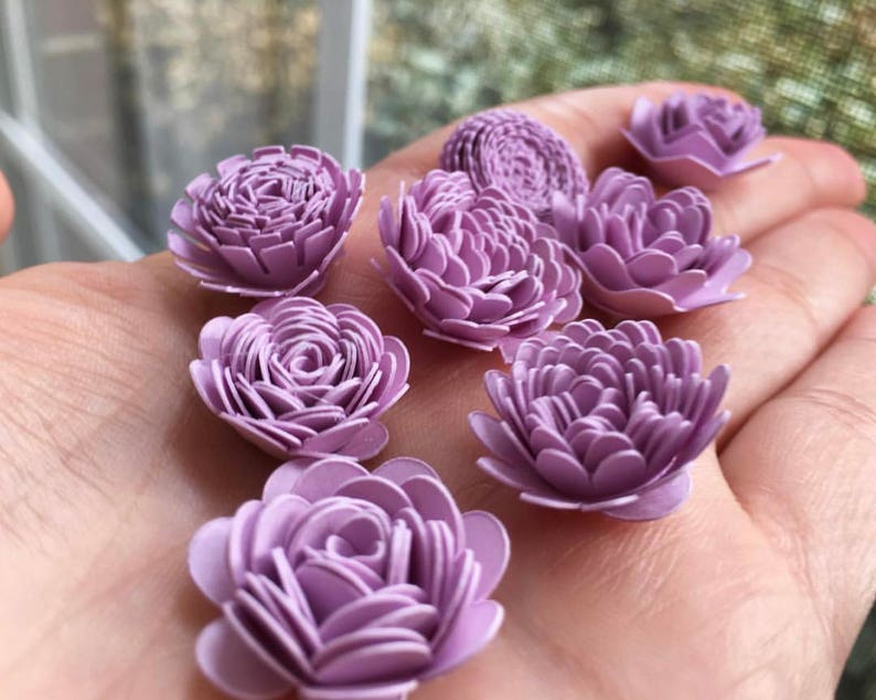 May include: A collection of lavender paper flowers, each with a different size and shape. The flowers are arranged on a hand, showcasing their delicate details.