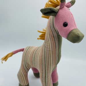 May include: A striped pink and green stuffed giraffe toy with a yellow mane. The giraffe has black eyes and a pink nose.