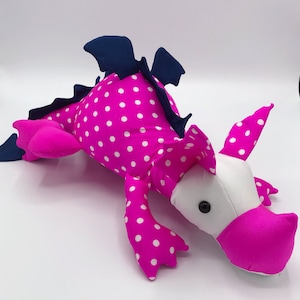 May include: Pink and white polka dot dragon plush toy with blue wings and a white face. The dragon has a pink snout and black eyes.