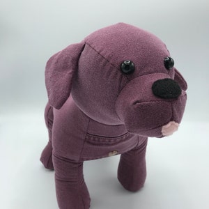 May include: A plush purple dog toy with black eyes and a black nose. The dog is wearing a denim vest.