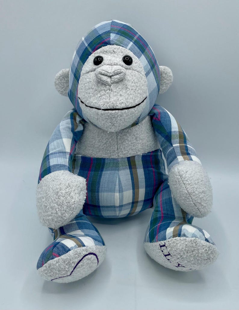 Puede incluir: Un mono de peluche azul y blanco a cuadros con pelaje gris. El mono tiene ojos negros y una sonrisa cosida. El juguete est&aacute; sentado sobre una superficie blanca.