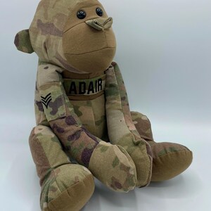 Puede incluir: Un mono de peluche con estampado de camuflaje y ojos de bot&oacute;n negros. El mono lleva una etiqueta con el nombre "ADAIR" y un parche de rango militar.
