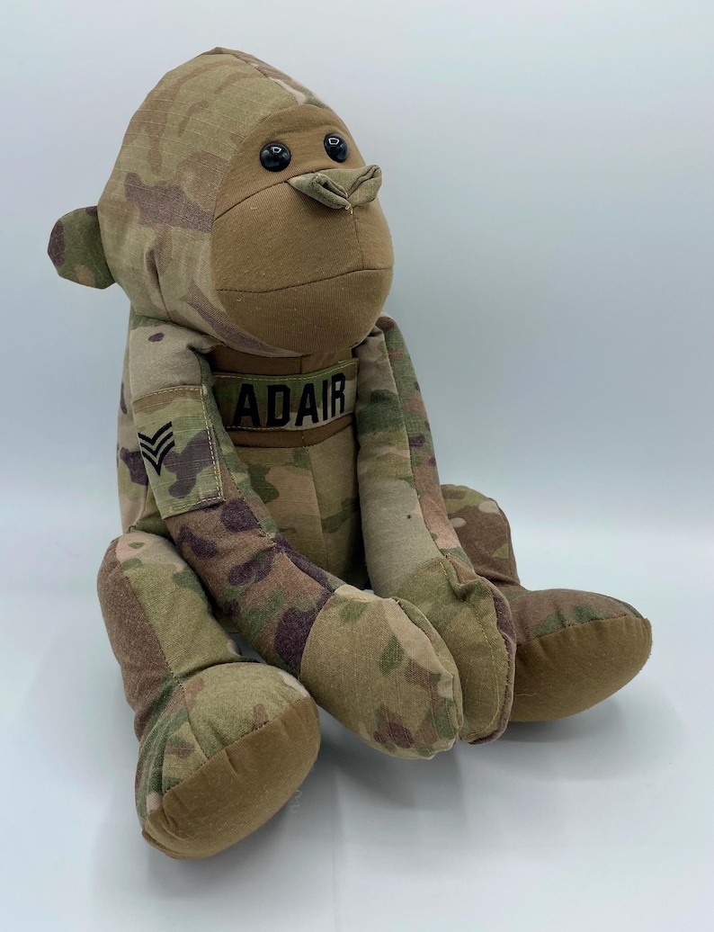 Puede incluir: Un mono de peluche con estampado de camuflaje y ojos de bot&oacute;n negros. El mono lleva una etiqueta con el nombre "ADAIR" y un parche de rango militar.