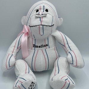 Puede incluir: Un mono de peluche blanco, rosa y azul a rayas con una cinta rosa alrededor del cuello. El mono est&aacute; bordado con el nombre "Emelina".