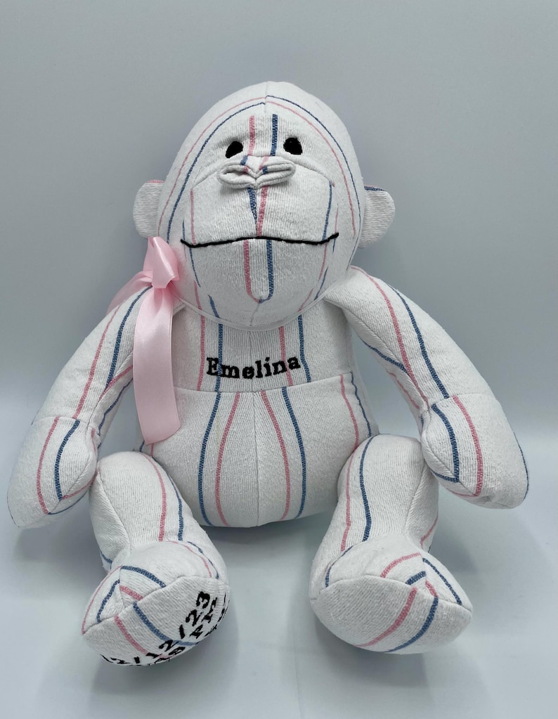 Puede incluir: Un mono de peluche blanco, rosa y azul a rayas con una cinta rosa alrededor del cuello. El mono est&aacute; bordado con el nombre "Emelina".