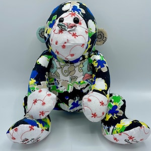 Puede incluir: Un juguete de peluche de mono de patchwork colorido con tela negra, blanca, azul, verde y amarilla. El mono est&aacute; sentado con las piernas extendidas y tiene una cara blanca con ojos negros y una boca roja.