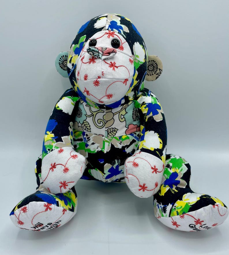 Puede incluir: Un juguete de peluche de mono de patchwork colorido con tela negra, blanca, azul, verde y amarilla. El mono est&aacute; sentado con las piernas extendidas y tiene una cara blanca con ojos negros y una boca roja.