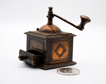 May include: A small, antique coffee grinder with a bronze finish. The grinder has a square base with a drawer, a hand crank, and a decorative diamond pattern. A coin is next to the grinder. The grinder is likely made of metal.