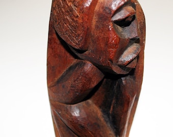 May include: A carved wooden sculpture of a human figure in a seated position. The sculpture is a rich brown color with visible wood grain and carved details. The figure's head is tilted downward, and the facial features are clearly defined.