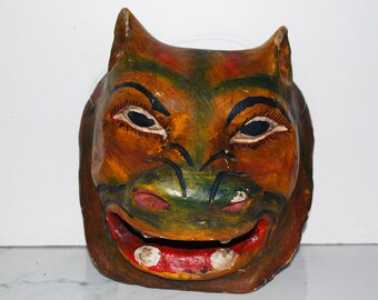 May include: A hand-painted wooden mask with a smiling face. The mask features a brown, orange, and green color scheme with black accents around the eyes and mouth. The mask has two small ears and a wide, red-lined mouth.