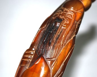 May include: A carved wooden figurine depicting an elderly man with a long beard. The figure is a warm brown color with darker accents, and the carving details are intricate. The figurine appears to be part of a larger object.