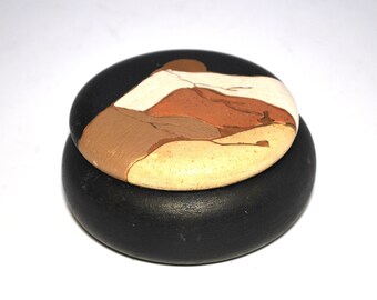 May include: A black, round container with a lid. The lid is decorated with a landscape design in shades of brown, tan, and white. The container is likely used for storage or display.