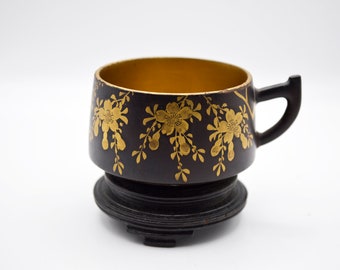 May include: A black teacup with a gold interior and a matching saucer. The cup is adorned with gold floral designs. The handle is black and the cup is approximately 3 inches tall.