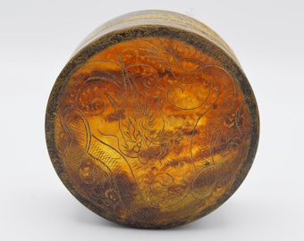 May include: A cylindrical container with a detailed dragon design etched into the top. The container has a warm, amber-colored interior and a dark brown exterior. The dragon is the focal point of the design.