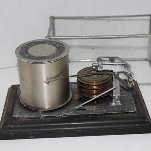 May include: An antique barometer with a cylindrical metal component, a series of copper-colored coils, and a small glass enclosure. The device is mounted on a dark wooden base and features intricate mechanical details.