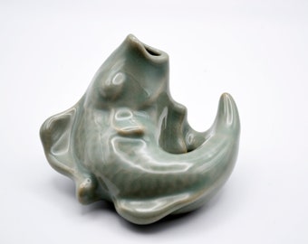 May include: A celadon-glazed ceramic fish-shaped vase. The vase has a curved body with a fin-like handle and a spout at the top. The fish is a light green color and has a glossy finish. The vase is likely for decorative purposes.