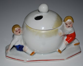 May include: A white ceramic ashtray with a round, lidded bowl and two small figurines of children. The children are wearing white shirts, blue shorts, and red shorts. The ashtray has a red border.
