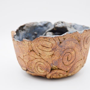 Könnte beinhalten: Eine handgefertigte Keramikschale mit einem einzigartigen, strukturierten Design. Die Schale hat eine braune Außenseite mit Spiralmustern und eine dunkelblaue Innenseite. Der Rand hat eine unebene, organische Form. Diese dekorative Schale ist ideal für die Inneneinrichtung.