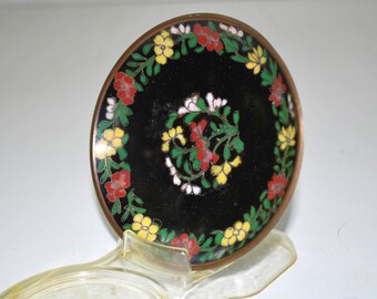 May include: A round, black decorative dish with a floral design. The dish has a bronze-colored rim and is decorated with red, yellow, and white flowers and green leaves. The dish is supported by a clear plastic stand.