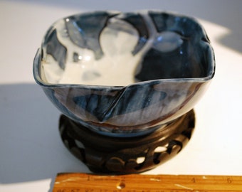 May include: A square ceramic bowl with a blue and white abstract design, resting on a dark wooden stand. The bowl has a unique shape with a slightly irregular rim. The stand is intricately carved.