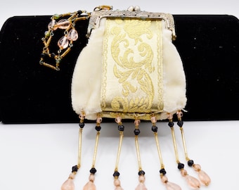 May include: Ivory-colored vintage purse with a gold floral design on the front. The purse has a metal clasp and is adorned with beaded fringe in black and pink. A beaded chain is attached to the purse.