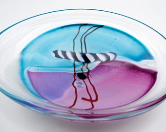 May include: A circular glass plate featuring a clear rim and a central design of swirling blue and purple pigments. The center includes a horizontal black and white striped glass element accented by three thin, vertical dark brown glass lines.