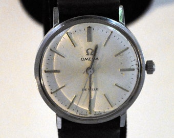May include: A vintage Omega De Ville wristwatch with a silver-colored face and a black leather band. The watch face features silver-colored hour markers and hands, with the Omega logo and "De Ville" text. The watch has a silver-colored case and a crown on the side.