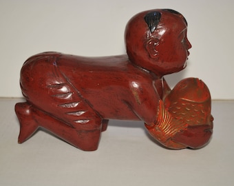 May include: A red wooden sculpture of a child crawling and holding a fish. The child's hair is black, and the fish has gold accents. The sculpture is on a white surface.
