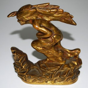 May include: A gold-colored sculpture of a winged figure in a dynamic pose, possibly a mythological being. The figure is perched on a textured base, with intricate details in the wings and clothing. The sculpture has a metallic finish.