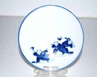May include: A white ceramic plate with a blue rim and a blue illustration of three figures playing. The figures are dressed in traditional clothing. A butterfly is also depicted. The plate is round.