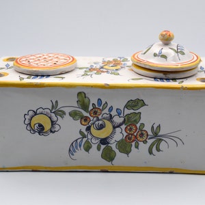 May include: A rectangular, white ceramic inkwell with a yellow border and floral design. The inkwell has a lid and a small, round, perforated cover. The floral design includes yellow, orange, and blue flowers.