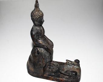 May include: A bronze-colored Buddha statue in a seated position, with a detailed head and a pointed topknot. The statue is on a rectangular base, with a smaller figure lying down in front of the Buddha. The statue has a weathered appearance.