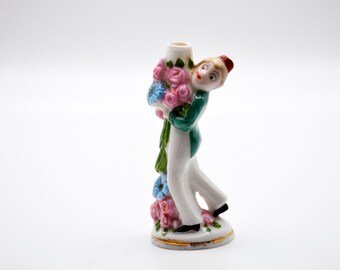 Może przedstawiać: Ceramiczna figurka osoby niosącej kompozycję kwiatową. Postać ubrana jest w zieloną kurtkę, białe spodnie i czerwony kapelusz. Kwiaty są różowe i niebieskie, z zielonymi liśćmi. Podstawa jest biała ze złotym obramowaniem.