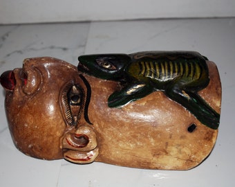 May include: A carved wooden sculpture of a face with a frog on top. The face is painted brown with red accents on the nose and mouth. The frog is green and black with yellow stripes.