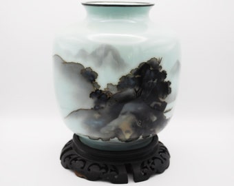 May include: A pale blue vase with a landscape painting of mountains and trees, set on a dark wooden stand. The vase has a rounded shape with a black rim and a detailed, artistic design.