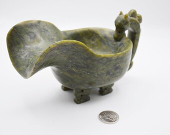 May include: A green and gray stone bowl with a unique, curved design. The bowl has three small feet and a handle with two decorative figures. A coin is next to the bowl for size comparison.