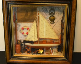 May include: A nautical-themed shadow box with a wooden frame. Inside, a miniature sailboat with white sails and a brown hull is the focal point. Other elements include a life preserver, a brass lantern, and a map background. The frame is 12 inches wide and 10 inches tall.