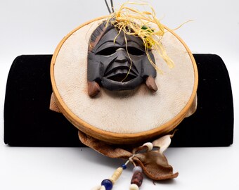 May include: A round, cream-colored decorative piece with a carved wooden mask. The mask is dark brown with a detailed face and is attached to a circular frame. The piece is adorned with dried grass, beads, and a seashell.
