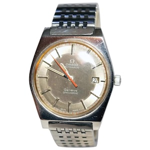 May include: A silver-toned Omega Genève Dynamic wristwatch with a gray face and a date display at the 3 o'clock position. The watch features a silver band and red second hand. The watch face has the words "OMEGA AUTOMATIC Genève DYNAMIC" printed on it.