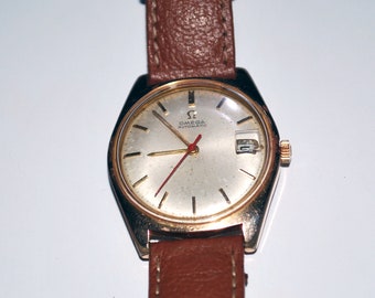 May include: A vintage gold-toned Omega Automatic wristwatch with a brown leather strap. The watch face is silver with gold hands and a red second hand. The watch has a date display at the 3 o'clock position.