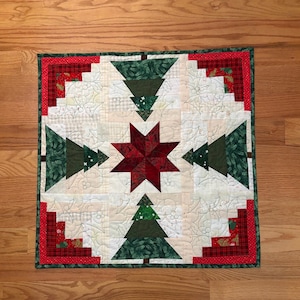 May include: A square quilted wall hanging featuring a central dark red star surrounded by four dark green pine tree shapes. The background consists of cream-colored fabric with stitched holly leaf patterns, framed by a red and green plaid border.