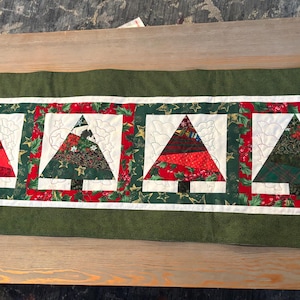 May include: A rectangular Christmas table runner featuring four stylized Christmas trees. Each tree is a patchwork of red, green, and white fabrics, set against a white background and framed by a red and green border. The runner has a green border.